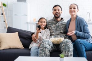 Smiling family with popcorn watching movie near father in camouflage uniform at home.